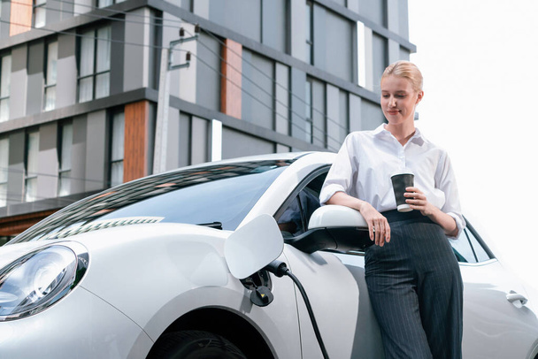 Businesswoman drinking coffee, leaning on electric vehicle recharging at public charging station with residential apartment condos building in background as progressive lifestyle by eco-friendly car. - Foto, Imagem