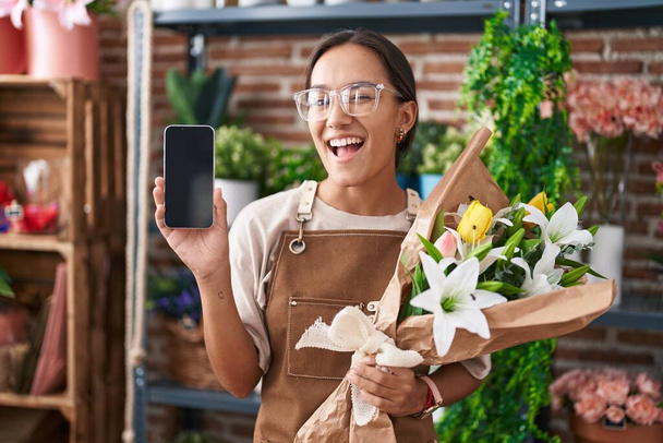 Giovane donna ispanica che lavora al negozio di fioristi che mostra lo schermo dello smartphone strizzando l'occhio alla fotocamera con espressione sexy, viso allegro e felice.  - Foto, immagini