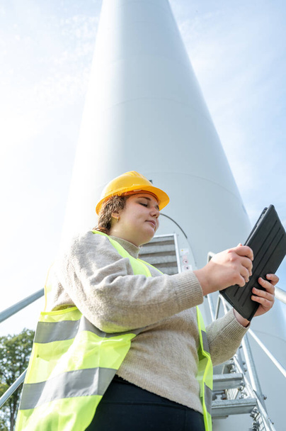Female engineer with brown curly hair and yellow helmet using her digital tablet while standing in front of a wind turbine during daylight, low angle view - Foto, afbeelding