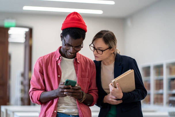 Ικανοποιημένος χαμογελαστός Αφροαμερικανός φοιτητής δείχνουν σοβαρή σκεπτική καυκάσιος συμμαθητής κορίτσι σε γυαλιά με εγχειρίδια στην οθόνη smartphone νέα δημιουργήθηκε app, viral δίκτυο βίντεο, διάλειμμα μελέτη αστεία θέση - Φωτογραφία, εικόνα