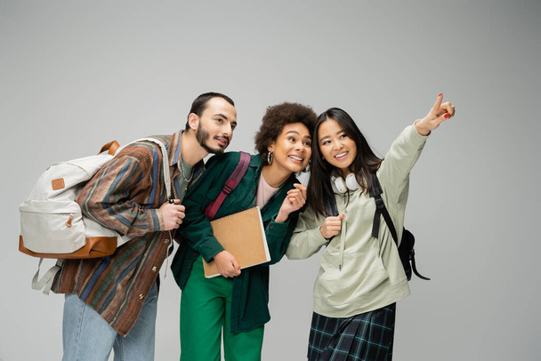 cheerful asian woman looking away and pointing with finger near multiethnic students isolated on grey - Foto, Bild