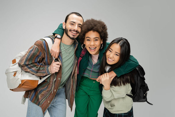 excited african american woman hugging young friends with backpacks isolated on grey - Foto, afbeelding