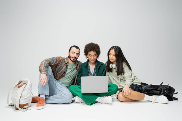 повна довжина молодої афроамериканки сидить з ноутбуком поруч зі стильними міжрасовими друзями на сірому фоні
 - Фото, зображення