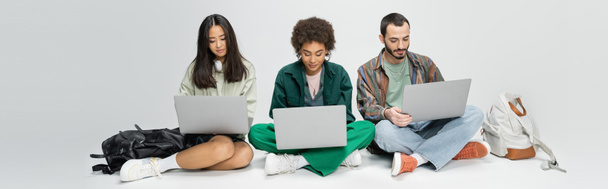 longitud completa de los estudiantes multiétnicos con estilo utilizando computadoras portátiles mientras se sienta cerca de las mochilas sobre fondo gris, pancarta - Foto, imagen
