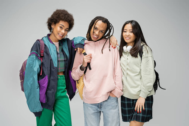 pretty multicultural women posing near trendy man with dreadlocks and backpack isolated on grey - Fotografie, Obrázek