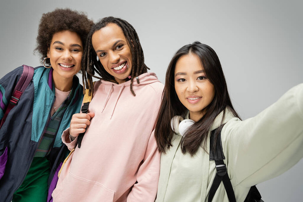 estudiantes multiculturales alegres en sudaderas con capucha y chaqueta posando y mirando a la cámara aislada en gris - Foto, imagen