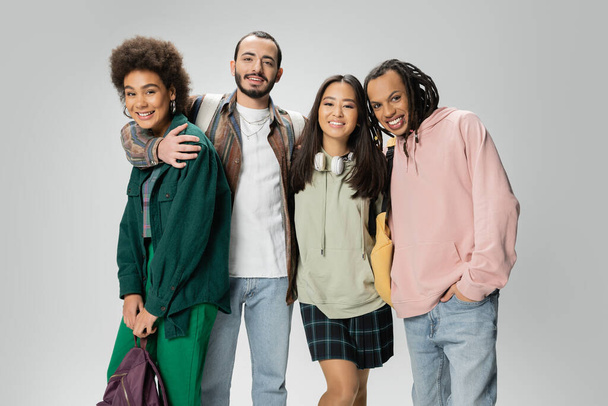 mosolygó multikulturális diákok divatos kapucnis pulóverben és ingben a kamerába néznek, miközben szürke hátizsákokkal állnak - Fotó, kép