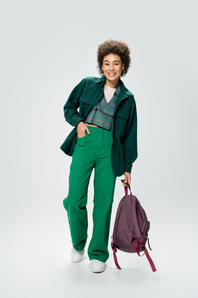 full length of stylish african american woman holding backpack while posing with hand in pocket of green pants on grey background - Foto, afbeelding