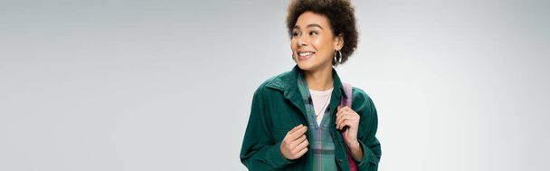 cheerful african american woman in stylish shirt standing with backpack and looking away isolated on grey, banner - Photo, image
