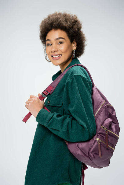 cheerful african american woman in shirt and hoop earrings holding backpack and looking at camera isolated on grey - Foto, immagini