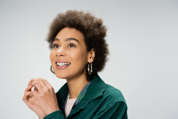 портрет взволнованной африканской американки с вьющимися брюнетками и серьгами на кольце, смотрящими в сторону изолированными на сером - Фото, изображение