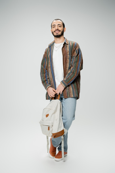 full length of cheerful bearded student in trendy outfit standing with backpack on grey background - Foto, Bild