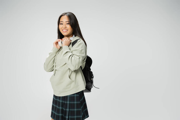 pleased asian woman in hoodie and plaid skirt posing with wireless headphones and backpack isolated on grey - Foto, imagen