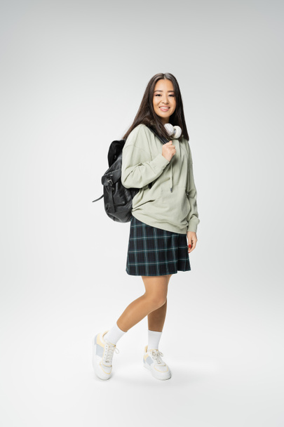 full length of cheerful asian woman with backpack and wireless headphones posing in plaid skirt and hoodie on grey background - Foto, afbeelding