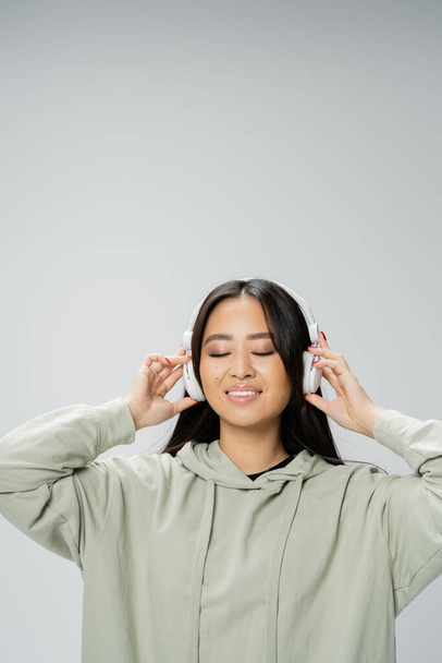 brunette asiatique femme en sweat à capuche écoute de la musique dans les écouteurs sans fil et souriant avec les yeux fermés isolé sur gris - Photo, image