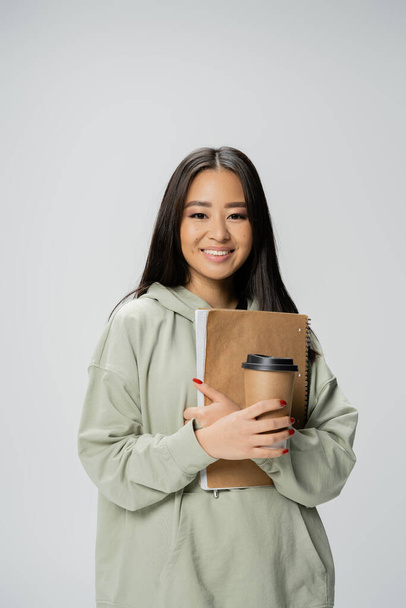 jovem asiático estudante no hoodie segurando takeaway bebida e copybook isolado no cinza - Foto, Imagem