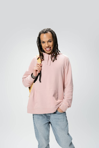 smiling multiracial student in pink hoodie holding backpack while posing with hand in pocket of blue jeans isolated on grey - Photo, Image