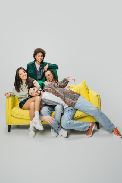joyful multicultural friends in trendy outfit having fun on yellow couch on grey background - Foto, Imagem