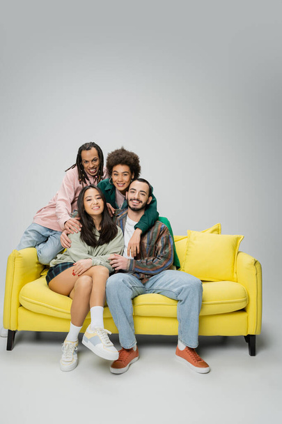 happy and stylish multicultural friends posing near yellow couch and looking at camera on grey background - Photo, image