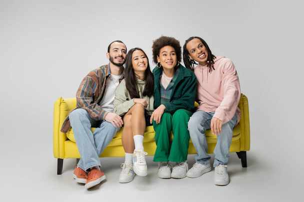 full length of smiling multicultural friends in trendy clothes watching movie on yellow sofa on grey background - Foto, immagini