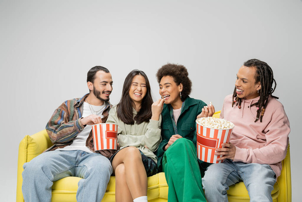 cheerful multiethnic friends in trendy clothes sitting with buckets of popcorn on yellow sofa isolated on grey - Photo, Image