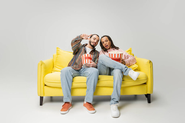 full length of bearded man pointing with finger while watching movie with multiracial friend with popcorn on grey background - Photo, Image