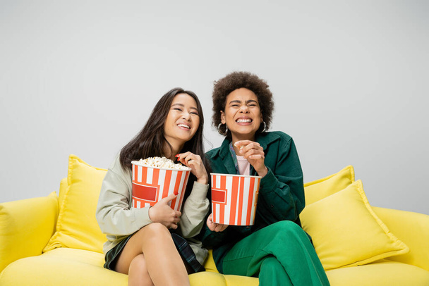 excited multiethnic women sitting on yellow couch with buckets of popcorn and watching movie isolated on grey - Photo, Image