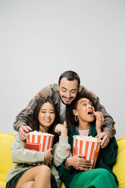 cheerful bearded man having fun while reaching popcorn near interracial women watching movie isolated on grey - Photo, Image