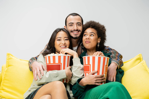 joyful bearded man hugging young multiethnic women sitting on yellow sofa with buckets of popcorn isolated on grey - Photo, Image