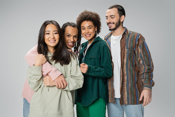 trendy multiracial man with dreadlocks embracing asian woman and smiling at camera near happy friends isolated on grey - Foto, Imagem