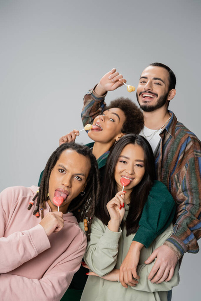 cheerful multiethnic friends in trendy clothes eating sweet lollipops isolated on grey - Foto, Imagen