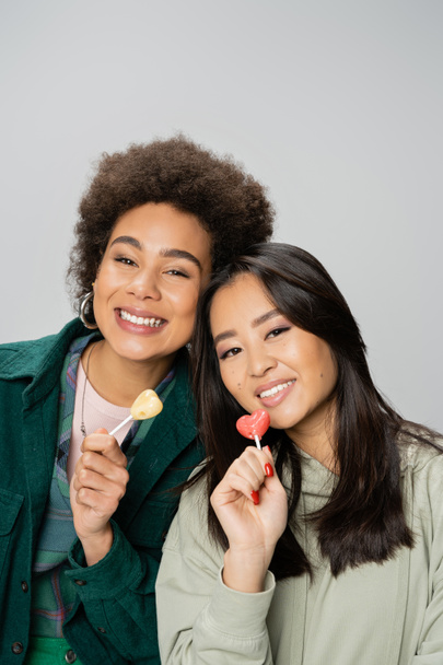 happy african american and asian women holding lollipops and looking at camera isolated on grey - Photo, Image
