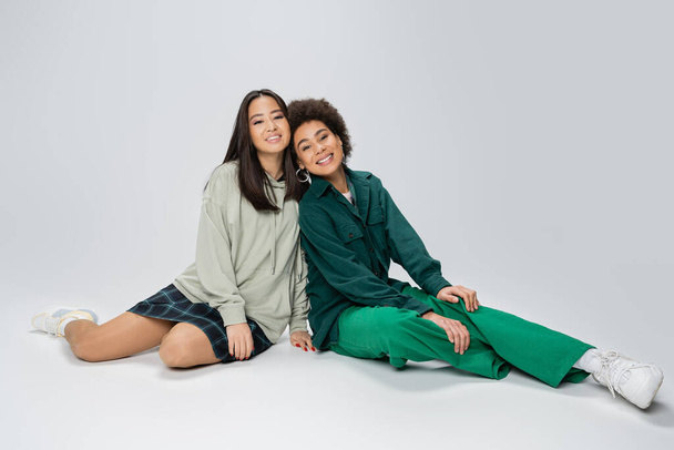 full length of asian and african american women in trendy outfit sitting and looking at camera on grey background - Photo, image