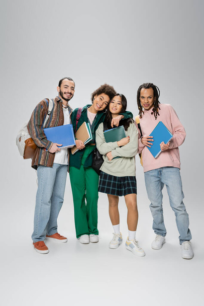 full length of happy multiethnic students with backpacks and notebooks looking at camera on grey background - Photo, Image