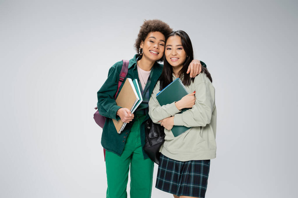 pretty interracial students with copybooks and backpacks smiling at camera isolated on grey - Foto, Imagem