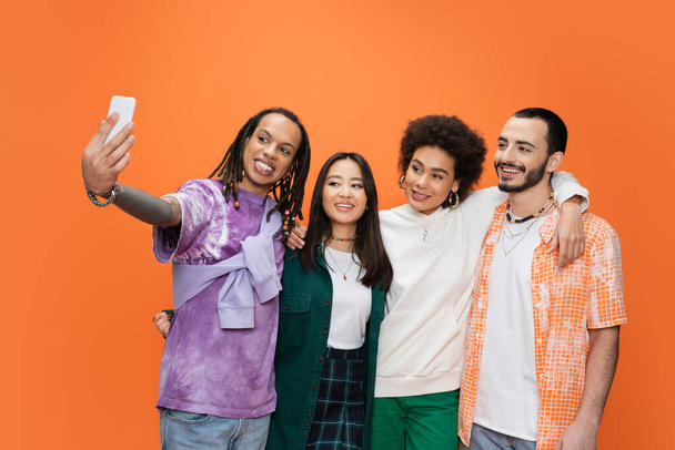 homem alegre com dreadlocks tomando selfie com jovens amigos multiculturais isolados em laranja - Foto, Imagem
