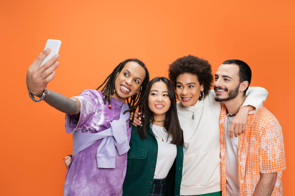 hombre de moda con rastas tomando selfie con amigos multiculturales felices aislados en naranja - Foto, Imagen