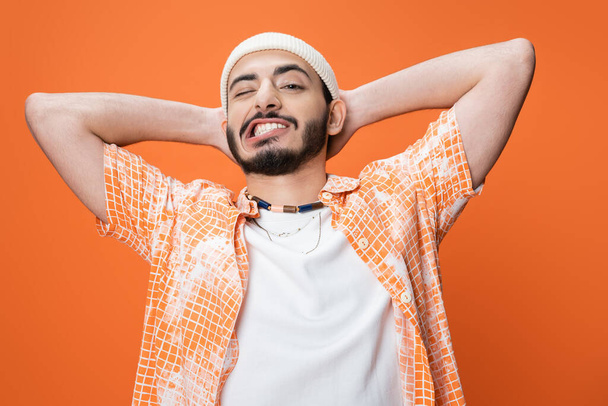 stylish bearded man in white beanie holding hands behind head and grimacing isolated on orange - Foto, Bild