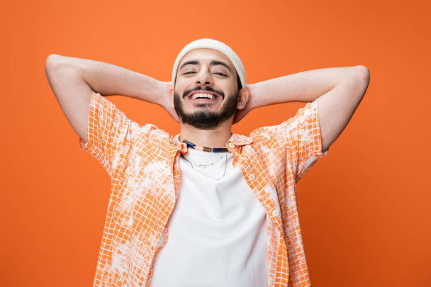 веселый бородатый мужчина в модном наряде позирует с руками за головой изолированы на оранжевый - Фото, изображение