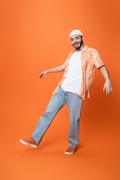 full length of cheerful bearded man in trendy shirt and blue jeans posing on orange background - Fotó, kép
