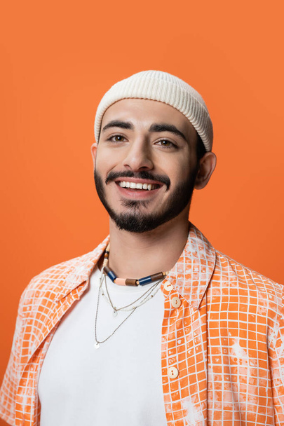 portrait of stylish bearded man in beanie and beads smiling at camera isolated on orange - 写真・画像