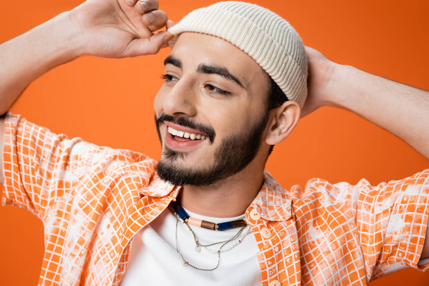 portrait of cheerful bearded man in beads touching white beanie and looking away isolated on orange - Photo, Image