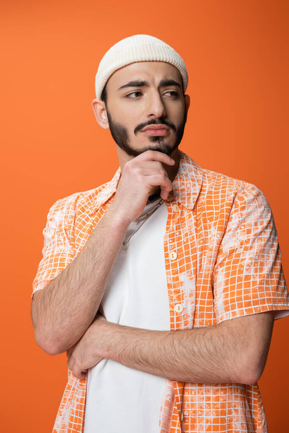 upset bearded man in stylish shirt and white beanie holding hand near chin and looking away isolated on orange - Foto, immagini