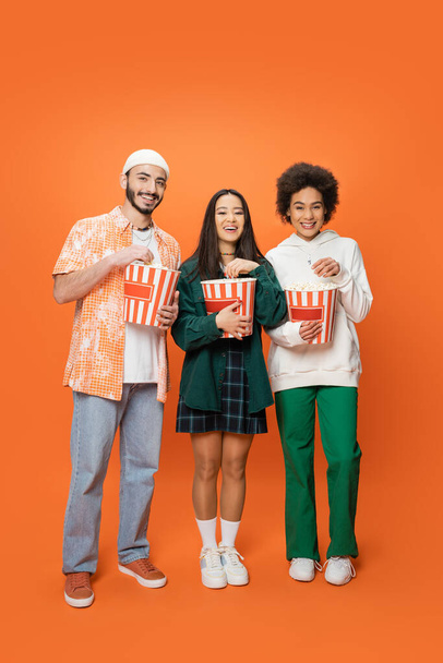 in voller Länge trendige multiethnische Freunde stehen mit Eimern Popcorn und lächeln in die Kamera auf orangefarbenem Hintergrund - Foto, Bild