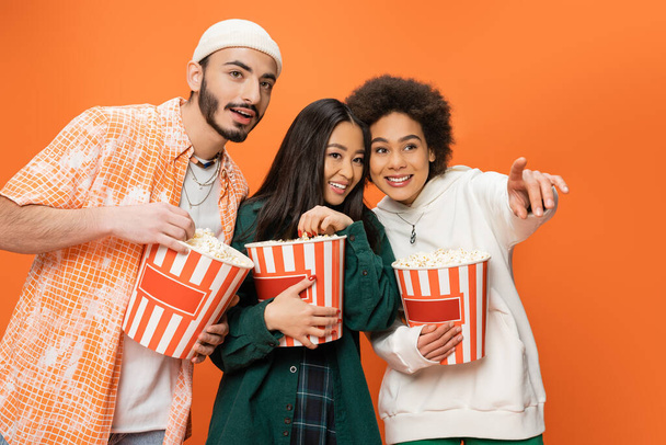 cheerful african american woman looking away and pointing with finger near multiethnic friends with popcorn isolated on orange - Foto, imagen