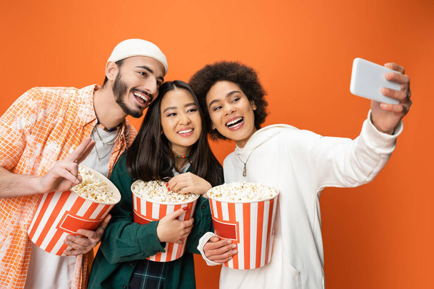 happy african american woman taking selfie with trendy friends holding buckets of popcorn isolated on orange - Foto, Imagen