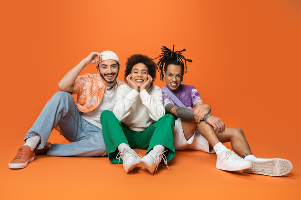 joyful multicultural friends in stylish outfit sitting and smiling at camera on orange background - Photo, Image