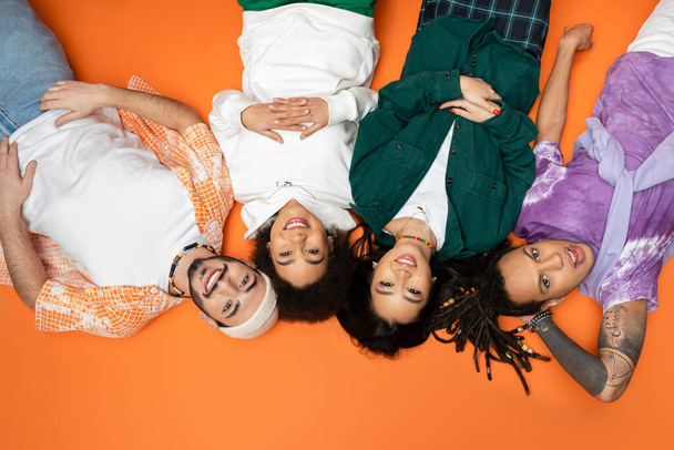 top view of joyful and stylish multicultural friends lying on orange background - Zdjęcie, obraz