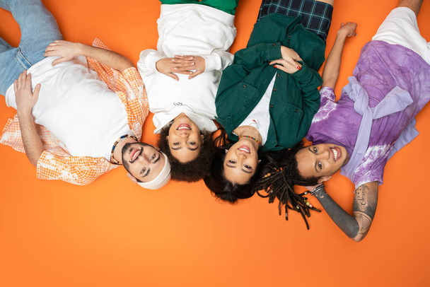 top view of happy multicultural friends in stylish outfit looking at camera while lying on orange background - Fotoğraf, Görsel
