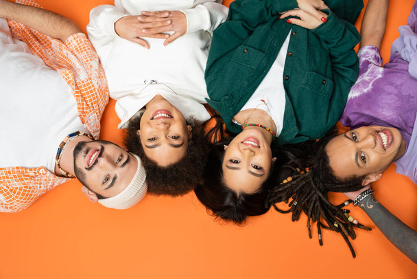 top view of pleased and trendy multiethnic friends lying and smiling at camera on orange background - Fotó, kép